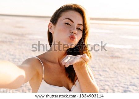 Picture of a cute happy young pretty girl walking outdoors at the beach and taking a selfie by camera while blowing kisses