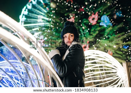 Happy woman in warm clothes poses in Christmas decorations at the fair, stands on the background of the Christmas tree and looks at the camera. Lady on Christmas Eve stands on a decorated street.