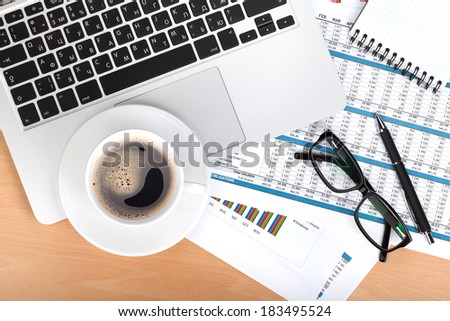 Coffee cup, laptop, notepad and glasses over papers with numbers and charts. View from above