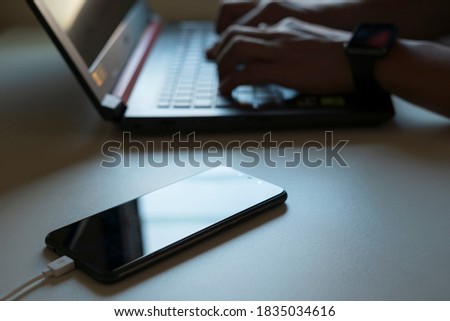 Close-up of a man working or typing on a laptop at a home desk with a battery-charged smartphone next to a warm light.