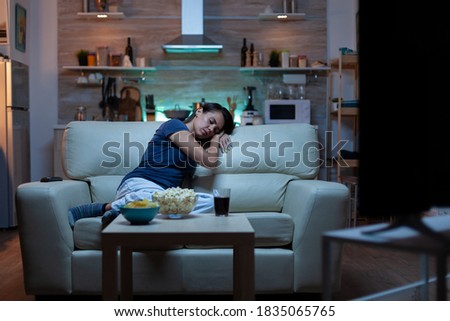 Young tired after work woman looking asleep in the evening in front of TV. Exhausted lonely sleepy lady in pajamas sleeping on sofa while watching a boring movie in living room, closing eyes at night