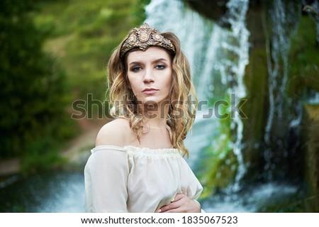 Young beautiful pregnant girl in a white dress and crown stands against the background of a waterfall 