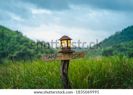 An old brown wooden sign with the words "TAKE IT EASY" on the lush green field. There was a square lamp with orange lights above the stump. Blurred background of trees and blue sky on cloudy days.