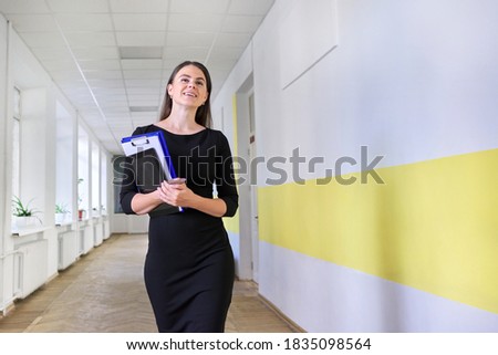 Portrait of young smiling female teacher at school. Female in black dress with folder, digital tablet walking along corridor