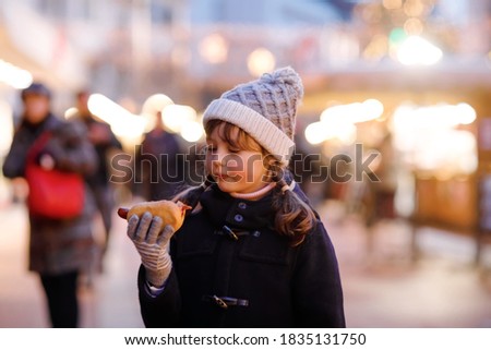 Cute little kid girl having fun on traditional Christmas market during strong snowfall. Happy child eating traditional curry sausage called wurst. schoolgirl standing by illuminated xmas tree.