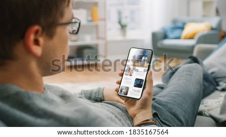 Over the Shoulder Shot of a Young Man at Home Sitting on a Sofa and Using Smartphone for Scrolling and Reading News about Technological Breakthroughs. He's Sitting On a Couch in His Cozy Living Room. Royalty-Free Stock Photo #1835167564
