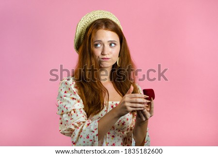 Disappointed upset girl, she doesn't like gift. Pretty european woman in floral dress holding small jewelry box with marriage proposal on pink background. 