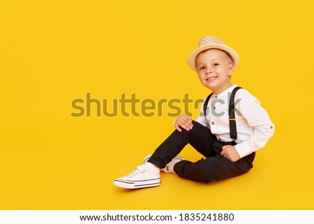 Stylish child boy in trendy black trousers with suspenders, white shirt and hat posing on yellow background