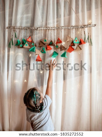 Cute kid opening handmade advent calendar with color paper triangles. Sweets hidden in colorful triangles hanging on branch. Seasonal activity for kids, family winter holidays, focus on calendar