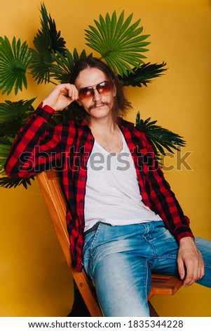 An attractive man with long hair and a mustache in a red plaid 80s disco shirt sits on a chair against . Guy in fashionable sunglasses on the background of a green palm tree