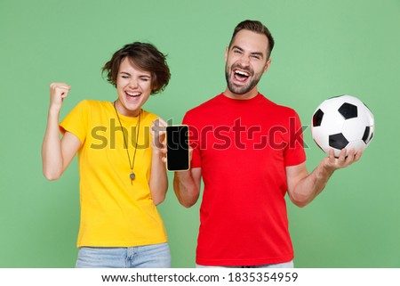 Happy couple friends sport family woman man football fans in t-shirt cheer up support team with soccer ball hold mobile phone with blank empty screen doing winner gesture isolated on green background