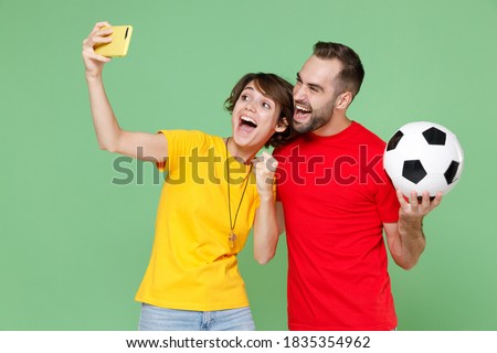 Excited couple friends sport family woman man football fans in t-shirts cheer up support favorite team with soccer ball doing selfie shot on mobile phone clenching fist isolated on green background