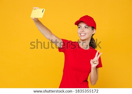 Delivery employee woman in red cap blank t-shirt uniform work courier in service during quarantine coronavirus covid-19 doing selfie shot on mobile phone isolated on yellow background studio portrait