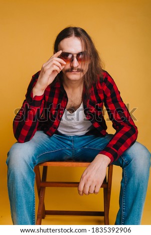 Attractive man with long hair and a mustache in a red 80s disco plaid shirt, sitting on a chair against a yellow background. Guy in fashionable sunglasses