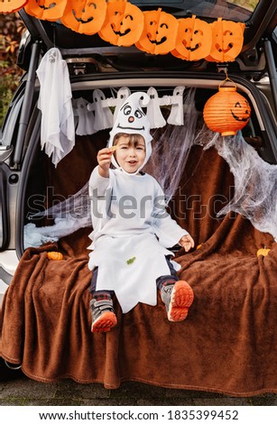 Trick or trunk. Trunk or treat. Happy child in ghost costume celebrating Halloween party in decorated trunk of car. New trend and alternative safe outdoor celebration of traditional holiday.