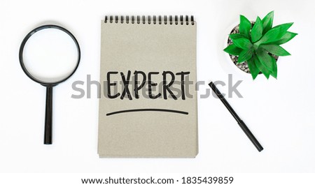 A magnifying glass, a blank brown notebook on a white table. EXPERT sign
