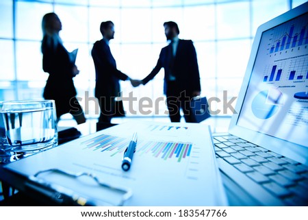 Laptop, financial document with pen and glass of water at workplace on background of three business partners making a deal Royalty-Free Stock Photo #183547766