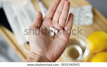 Supporting immune system in season of  flu or coronavirus. Vitamin D, vitamin C and zinc in senior hand on background of wooden table with face mask, sanitizer, thermometer and lemons Royalty-Free Stock Photo #1835540941