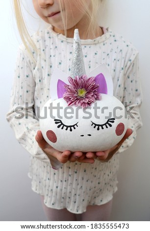 A girl holding a decorated pumpkin in the form of a unicorn with a horn and a flower wreath in her hands, autumn craft.