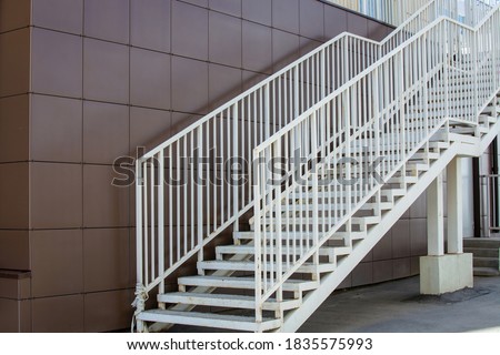 White wide metal pedestrian staircase with handrails at a city building with a brown tiled wall. Steel framed staircase for access to upper floor. Daylight urban background with place for copy space.