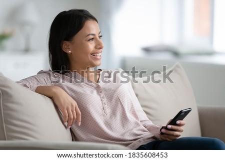 Smiling African American woman sit relax on couch at home use smartphone look in distance thinking dreaming. Happy biracial female rest on sofa in living room make plans or visualize future.