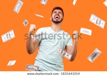 Money rain, winner and rich. Bottom view of ecstatic motivated man standing with raised fists and shouting for joy, winner excited for success. dollars falling. indoor isolated on orange background