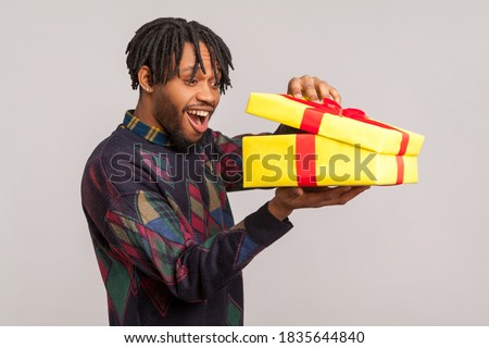 Profile portrait happy african man with dreadlocks opening gift box and peeping inside with toothy smile, satisfied with present. Indoor studio shot isolated on gray background Royalty-Free Stock Photo #1835644840