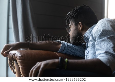 Upset young African American male sit on sofa at home look in distance mourning missing. Unhappy sad biracial guy distressed with life problems, feel lonely thinking or pondering. Solitude concept. Royalty-Free Stock Photo #1835655202