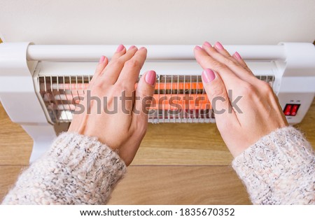 Woman warms her frozen hands near an infrared electric heater at home.  Royalty-Free Stock Photo #1835670352