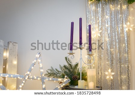 Christmas decorations. Purple candles in candlestick, fir tree branches and cones, siver curtains with shiny stars and warm garland lights and bokeh. 
