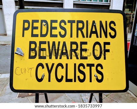 Yellow traffic sign reading 'pedestrians beware of cyclists' in London, UK. Someone has added 'cunt' in pen in front of cyclist, signalling a negative attitude to bicycles on British roads. 