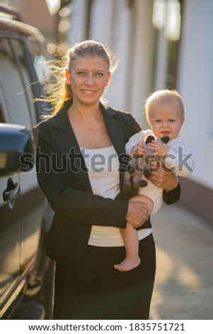 Beautiful business woman mother in a suit with a small child in her arms against the background of her house and a black car goes to work