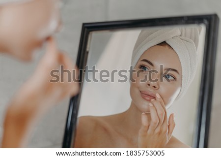 Healthy woman wears minimal makeup, takes care of complexion and lips, looks at herself in mirror, stands bare shoulders, wears bath towel on head, has healthy flawless skin. Beauty, hygiene concept Royalty-Free Stock Photo #1835753005