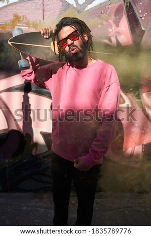 Excellent mood. Cheerful young artistic multiracial man with dreadlocks wearing sunglasses holding skateboard at his shoulder and posing to the camera with funny faces