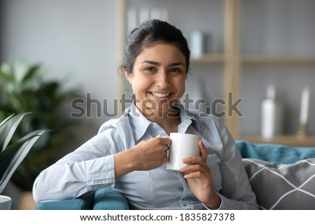 Pretty Indian girl sit on sofa with cup of fresh brewed tea relaxing alone in living room. Mixed-race housewife enjoy morning coffee at home feels happy, take break relish pause from housework concept