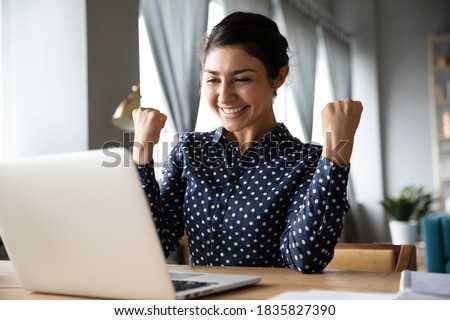 Overjoyed indian ethnicity girl sit at desk looks at laptop screen read incredible news clench fists makes yes gesture celebrate on-line lottery gambling win, getting new job offer feels happy concept Royalty-Free Stock Photo #1835827390