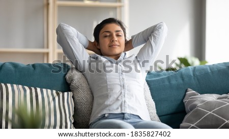 Serene Indian woman closed eyes breath fresh humified air in cozy living room alone put hands behind head lean on sofa having day nap alone, reduces fatigue, enjoy pleasant thoughts, no stress concept Royalty-Free Stock Photo #1835827402