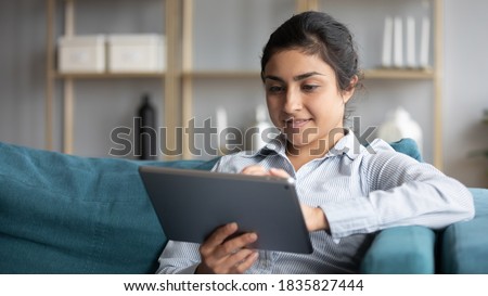 Calm indian woman resting on couch at home hold tablet modern device smile read message enjoy remote shopping online, distance chat with friend, plan vacation booking hotel and travel buy tour concept Royalty-Free Stock Photo #1835827444