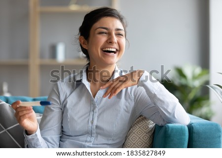 Happy Indian 30s female sit on sofa hold pregnancy positive two stripes test laughing enjoy news feels excited by pregnancy. Easy accurate method tool, healthy woman, expectant mother portrait concept Royalty-Free Stock Photo #1835827849