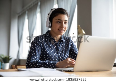 Indian business woman sit at desk wearing headphones lead negotiations through videoconference call with clients consulting customer use laptop. Educational webinar, on-line class distantly concept Royalty-Free Stock Photo #1835827852