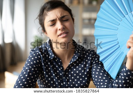 Overheated indian woman sit at workplace without air conditioner hold blue handheld fan cools herself frown face closed eyes feels unwell. Too hot summer weather day, high temperature indoors concept Royalty-Free Stock Photo #1835827864