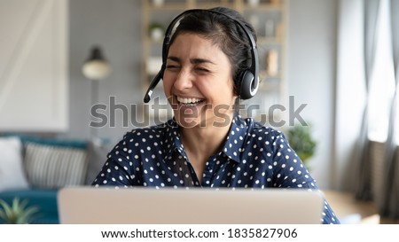 Cheerful Indian female wear headset with microphone sit in front of laptop laughing during video call communication. Distance e-learning, helpline work technical support happy worker portrait concept Royalty-Free Stock Photo #1835827906