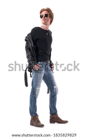 Side view of stylish handsome biker with red hair wearing sunglasses removing leather jacket. Full body isolated on white background.
