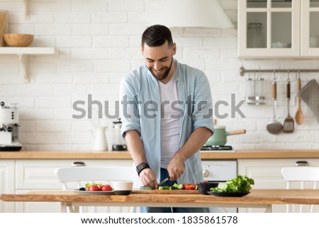 Smiling young bearded man cooking salad, cutting fresh vegetables in modern kitchen, happy satisfied male preparing vegetarian snacks, food for dinner or party, enjoying leisure time, hobby Royalty-Free Stock Photo #1835861578