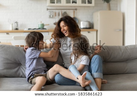 Overjoyed young mother having fun with two kids at home, laughing happy mum, adorable little daughter and son playing funny active game, tickling, sitting on cozy couch in living room Royalty-Free Stock Photo #1835861623