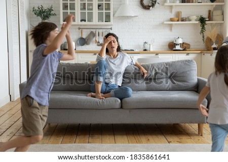 Exhausted tired mother touching forehead, sitting on couch, having problem with noisy hyperactive kids, naughty too active little son and daughter shouting and running around sofa at home Royalty-Free Stock Photo #1835861641
