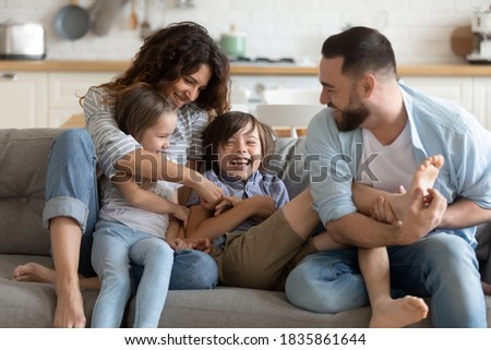 Happy young parents with two kids having fun at home, laughing overjoyed mother and father tickling little daughter and son, sitting on couch in living room, family enjoying leisure time together Royalty-Free Stock Photo #1835861644