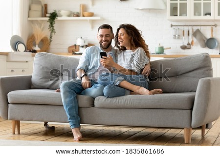 Happy young woman and man hugging, using smartphone together, sitting on cozy couch at home, smiling overjoyed wife and husband looking at phone screen, sitting on sofa in modern living room Royalty-Free Stock Photo #1835861686
