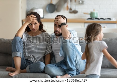 Tired mother and father having problem with noisy naughty daughter shouting and running around sofa at home, exhausted parents touching foreheads, sitting on couch, too active kid concept Royalty-Free Stock Photo #1835861719