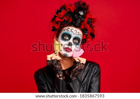 Girl with sugar skull makeup with a wreath of flowers on her head and skull, wearth lace gloves and leather jacket, keeps cosmetic sponge on eye isolated on red background.
